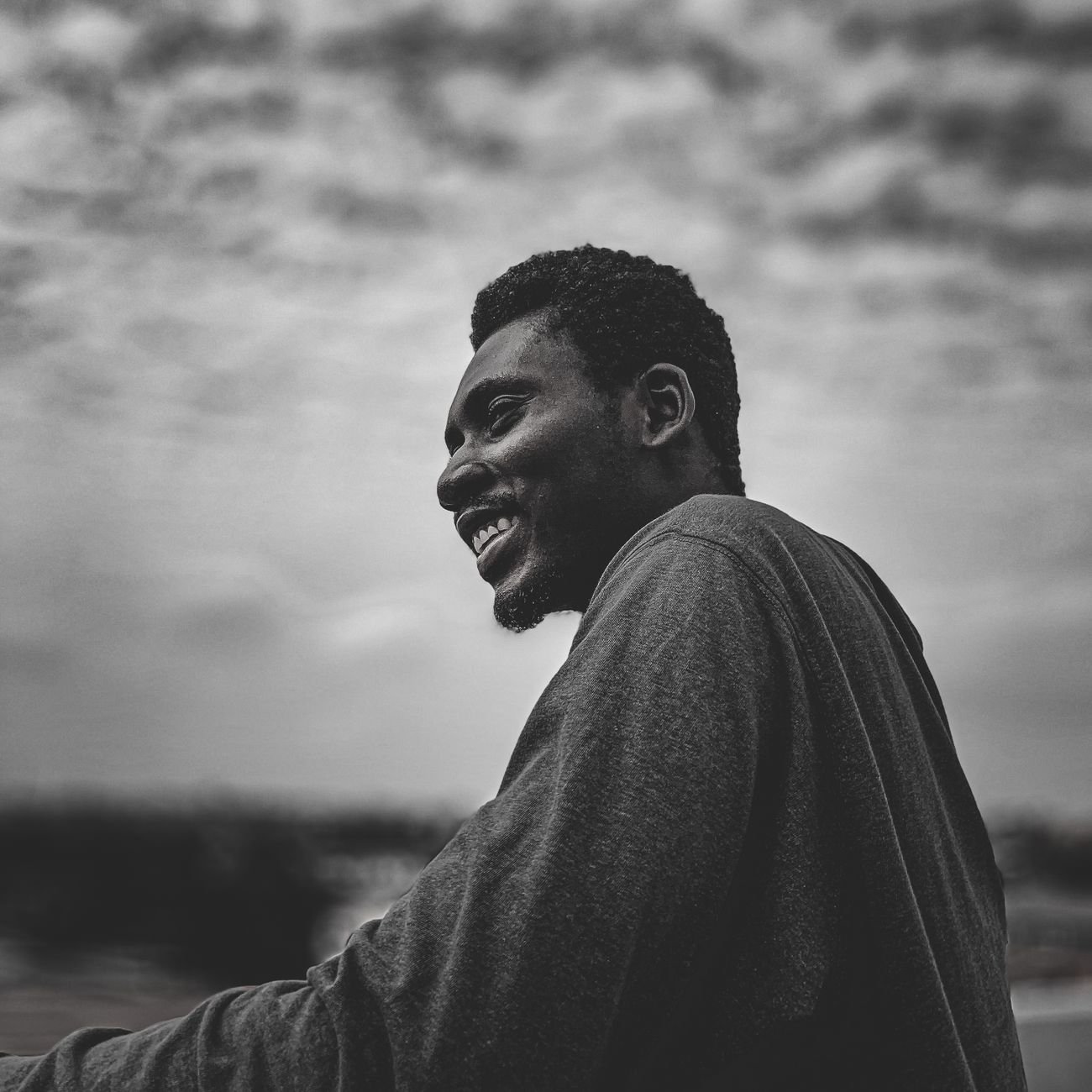 Black and white portrait of a smiling man outdoors in a cloudy setting.